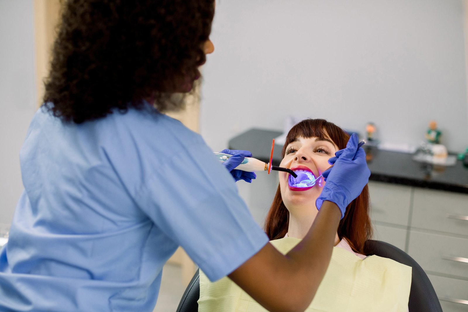 Back angle view of African woman dentist working with dental polymerization lamp in oral cavity of her female patient. Dental restoration with a photopolymer UV lamp. Dental treatment.